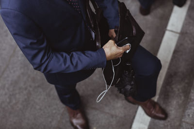 Low section of businessman with open bag holding smart phone and in-ear headphones in city