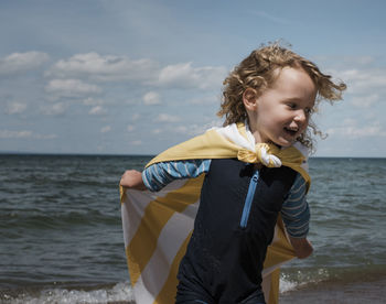 Happy girl wearing cape while running by sea against sky during sunny day