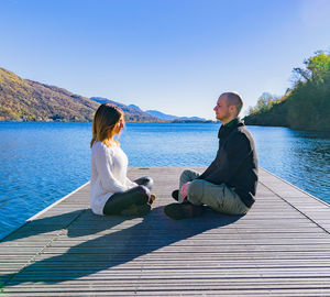 Man and woman sitting on pier in lake against clear sky