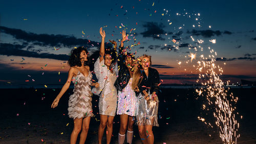 Cheerful friends holding confetti while standing against sky