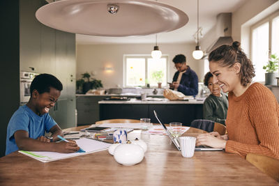 Happy mother using laptop with son coloring in book while sitting at dining table in home