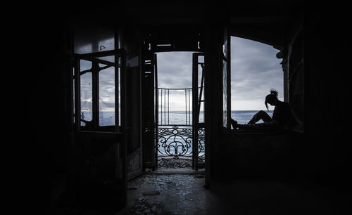 Silhouette woman sitting on window sill in abandoned home against sea