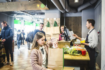 Daughter packing paper bag while parents and cashier at checkout counter in background