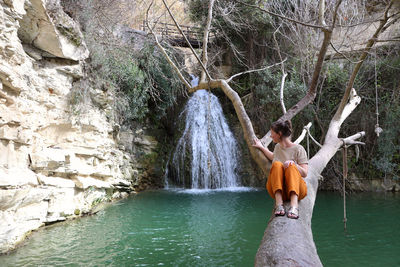 Woman looking at waterfall while sitting on tree trunk