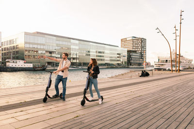 Couple exploring city while riding on electric push scooters at promenade by river