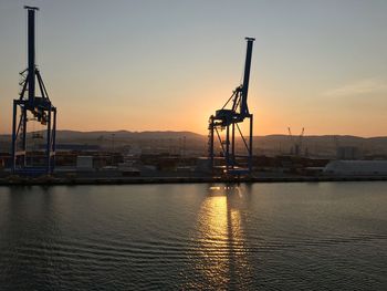 Cranes at harbor against sky during sunset