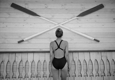 Rear view of woman in swimwear standing against oars mounted on wall