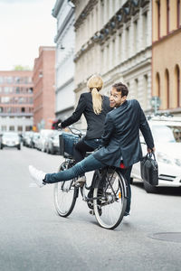 Rear view portrait of happy businessman sitting on back seat while female colleague riding bicycle