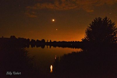 Silhouette trees by lake against sky at night