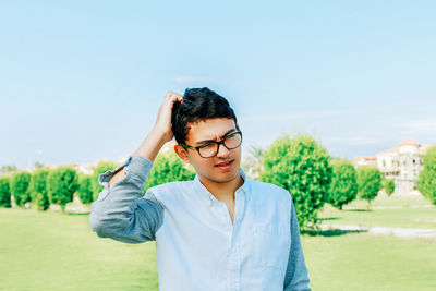 Young man scratching head while standing on field against sky