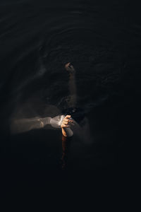 From above side view of unrecognizable female traveler covering face with textile while standing with raised arms in water at dusk