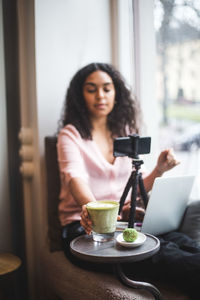 Young female influencer drinking matcha tea while making vlog through mobile phone by window at creative office