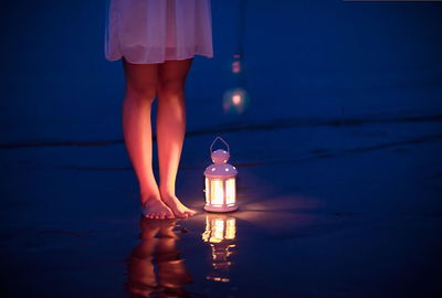 Low section of woman standing by illuminated sea at dusk