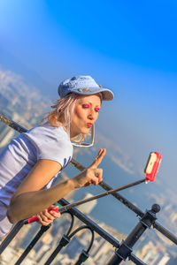 Smiling woman gesturing while taking selfie standing against railing in city