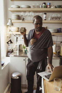 Businessman with son in baby carrier using laptop while holding cup at kitchen