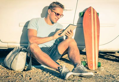 Man using digital tablet while sitting with skateboard against car