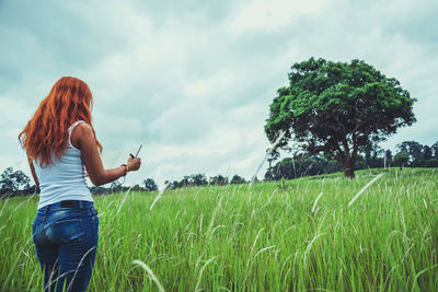 Rear view of woman reading book while standing on field against sky