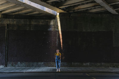 Young androgynous person waiting on sidewalk below bridge