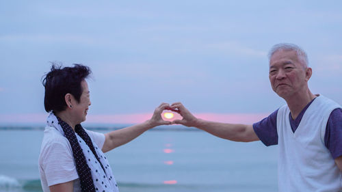 Optical illusion of man and woman holding dun in heart shape over sea against sky