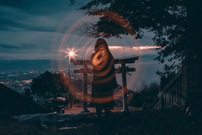 Rear view of woman standing by illuminated trees against sky at night