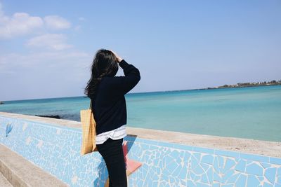 Side view of woman looking at sea while standing by retaining wall against blue sky during sunny day