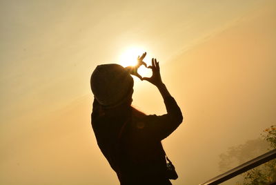 Silhouette woman making heart shape while standing against sky during sunset