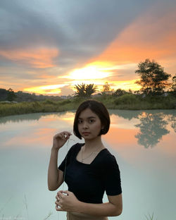 Portrait of young woman standing against lake during sunset
