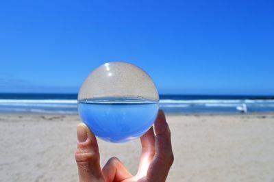 Cropped hand of person holding crystal ball on beach against clear blue sky