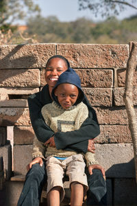 Portrait of smiling woman with son sitting against brick wall