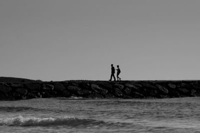 Silhouette friends walking on groyne by sea against clear sky