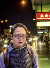 Portrait of young man standing on busy city road with illumination at night.