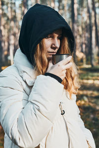 Woman in a hood having break during autumn trip holding cup with hot drink from thermos flask