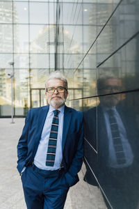 Senior business with hands in pockets leaning outside office building