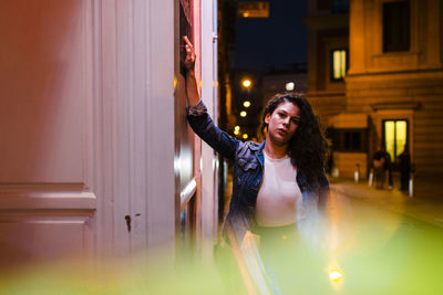 Confident young woman standing by wall on illuminated street at night