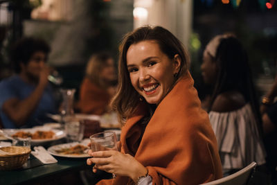 Portrait of smiling young woman holding drink while sitting at table during dinner party in backyard