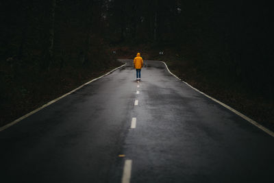 Back view of anonymous man in yellow jacket walking on empty asphalt road among green forest