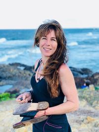 Portrait of smiling mature woman holding slippers while standing against sea at beach