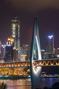 Illuminated bridge over river by buildings against sky at night