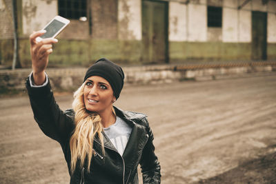 Fashionable young woman wearing leather jacket taking selfie with smart phone