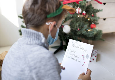 Rear view of boy wearing mask holding paper sitting against christmas tree at home