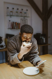 Man eating doughnut and using smart phone while leaning on table at home