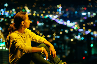 Side view of woman sitting on retaining wall against illuminated city at night