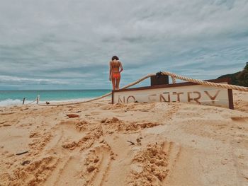 Woman in bikini walking on shore by no entry text at beach against cloudy sky