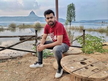 Portrait of young man sitting by lake against sky