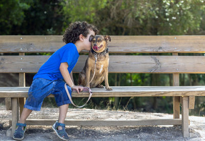 Child kissing his dog with love on a wooden bench in a natural park, on a sunny summer day,