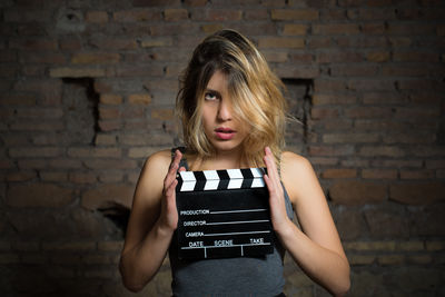 Portrait of young woman holding film slate against brick wall