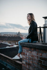 Young woman sitting on retaining wall against cityscape during sunset