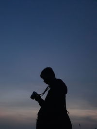 Silhouette woman holding camera against sky during sunset