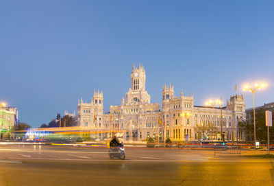 Beautiful cybele palace and illuminated city square with motorcyclist under evening blue sky in madrid