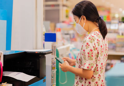 Cropped image of female in protective mask holding cell phone with printer to print the document.
